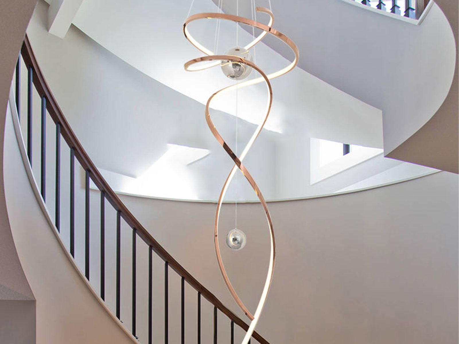 Modern rose gold stainless steel helical helix LED wave pendant light with floating orb spheres suspended in a high ceiling foyer